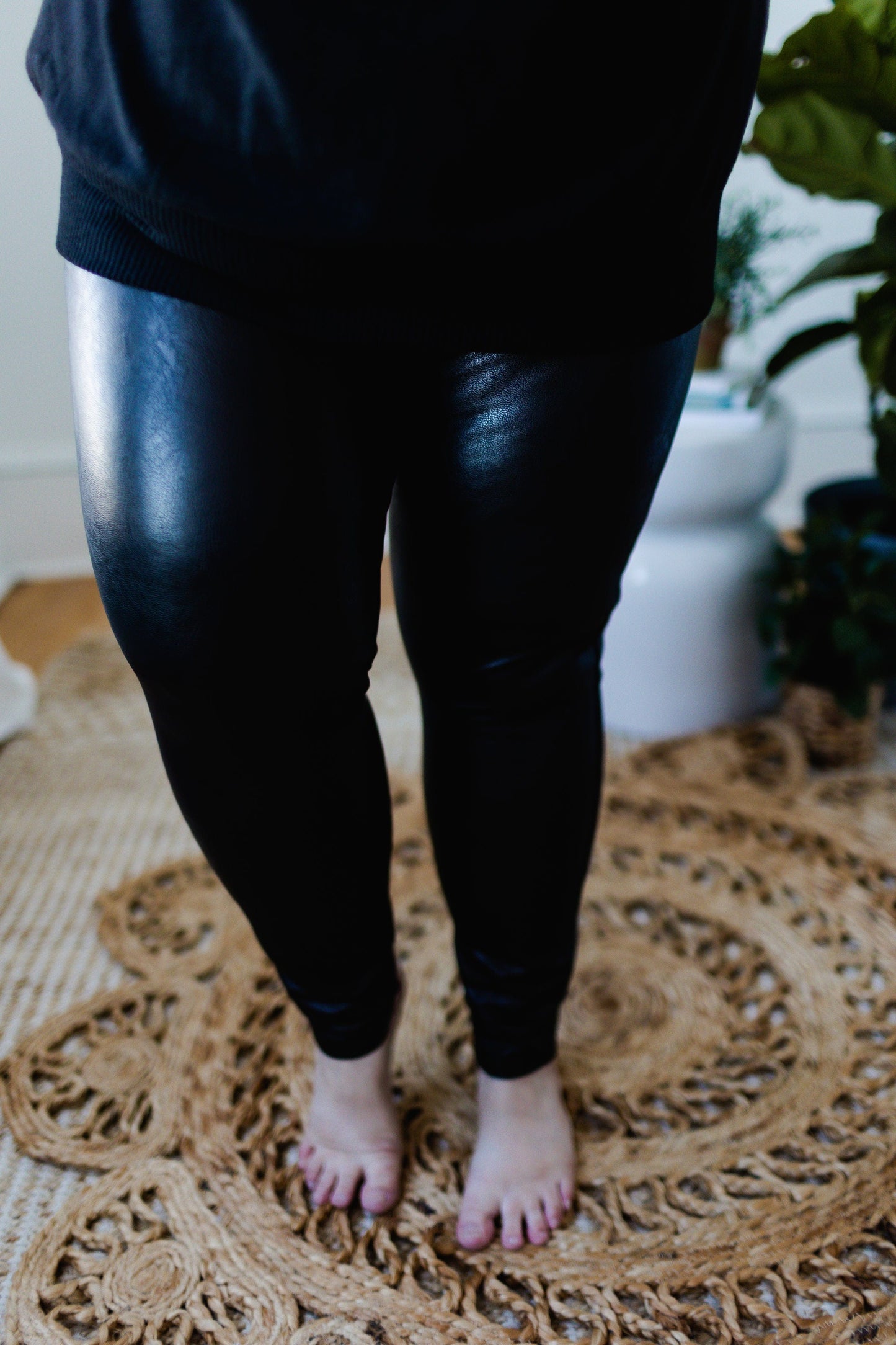 A person wearing Spanx© Leather-Like Ankle Skinny Pants stands barefoot on a woven circular rug, with plants in the background.