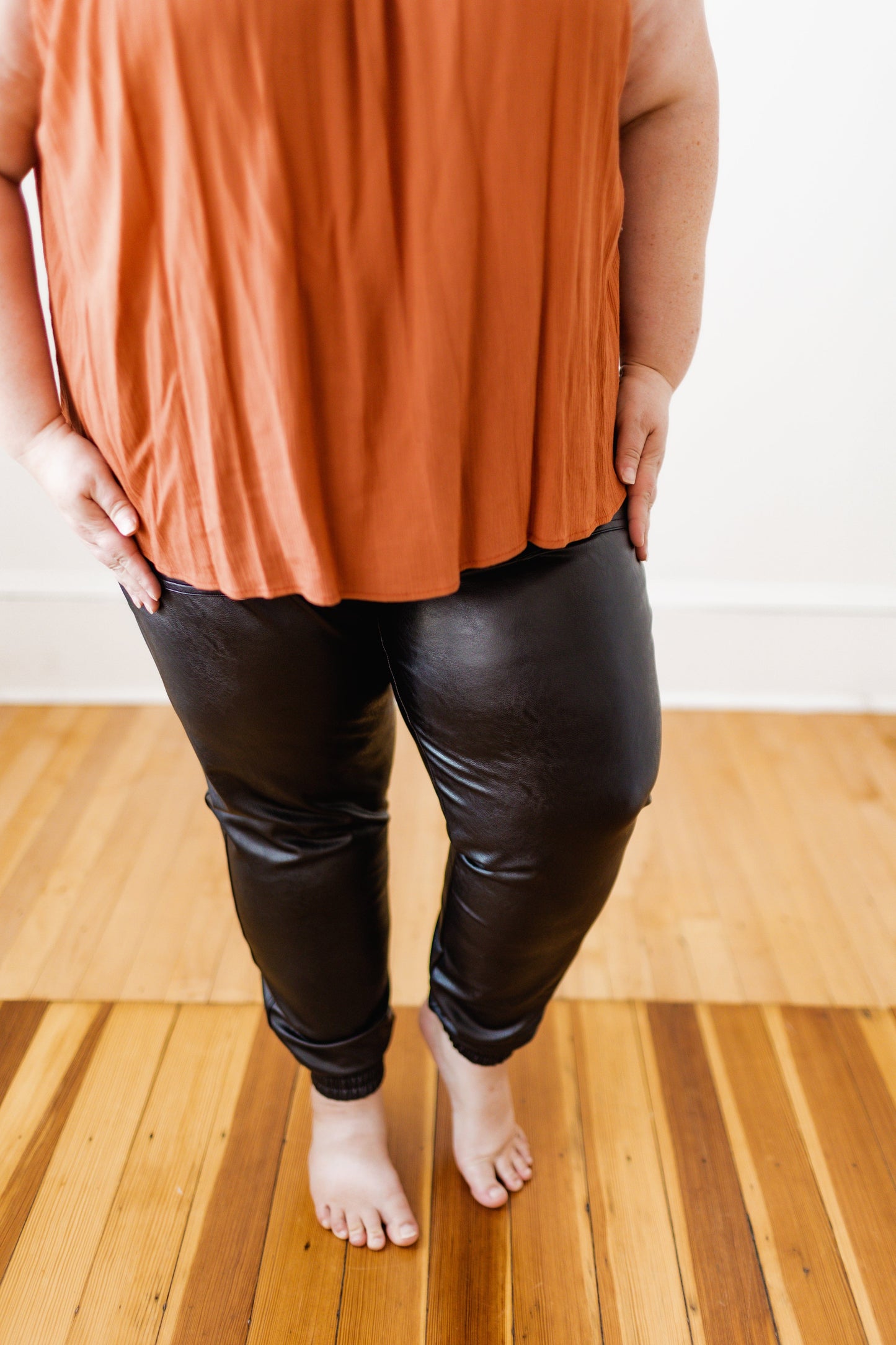 A person in a rust-colored top and comfortable Spanx© Leather-Like Jogger pants from Spanx stands barefoot on a wooden floor.