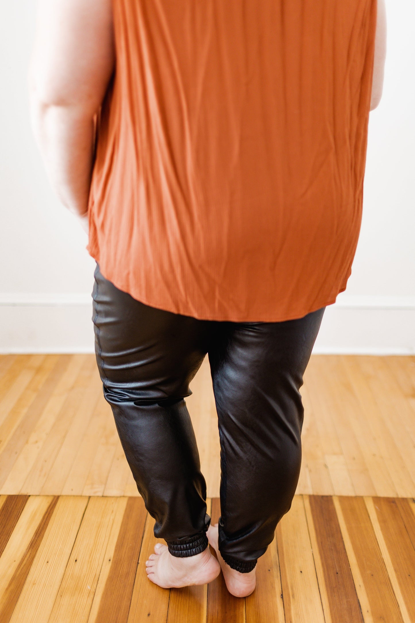 A person in a sleeveless orange top and Spanx© Leather-Like Jogger pants stands barefoot on a wooden floor, facing away.