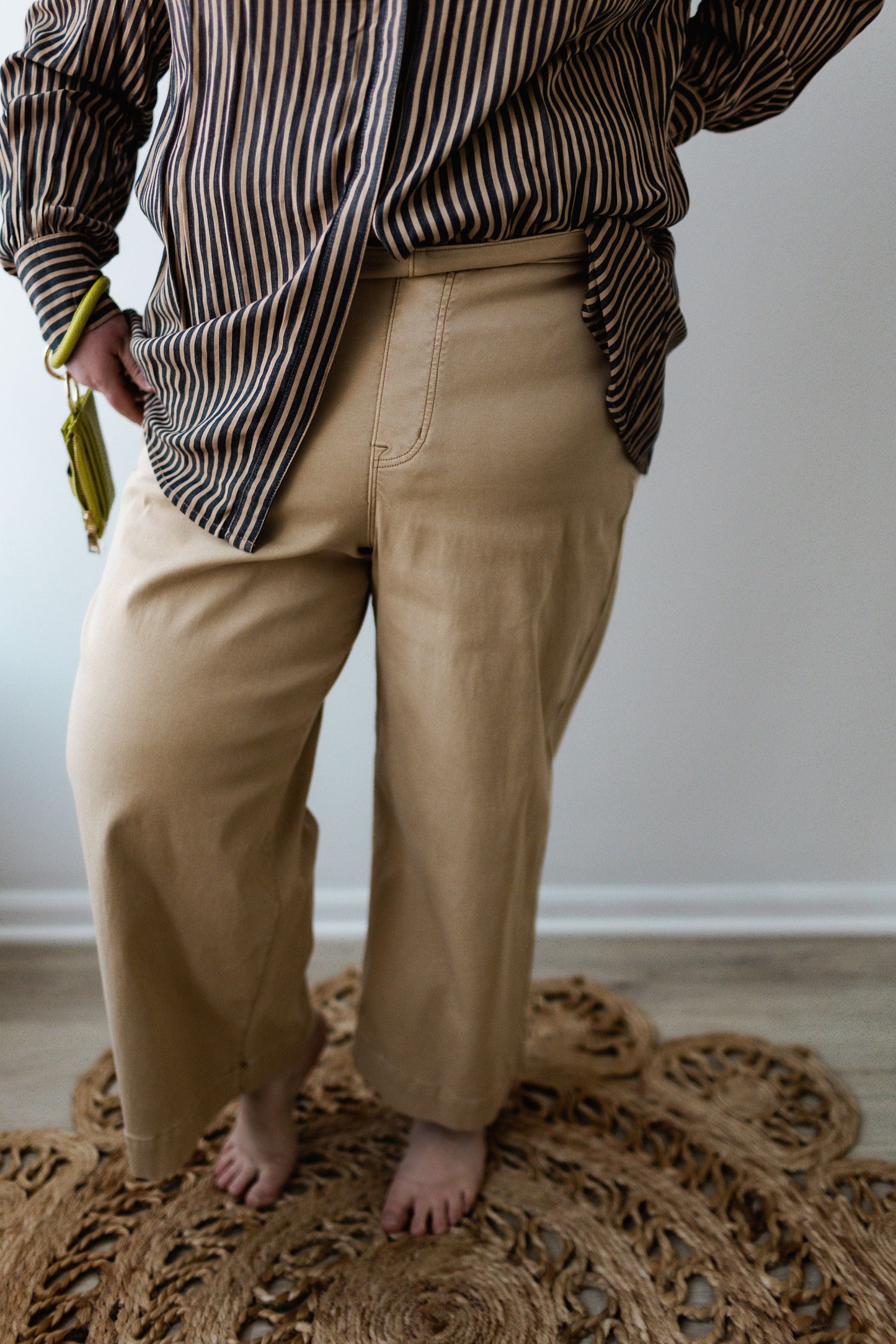 A person in a striped shirt and Spanx® STRETCH TWILL CROPPED WIDE LEG PANT IN ALMOND stands on a woven rug, holding a small yellow purse.