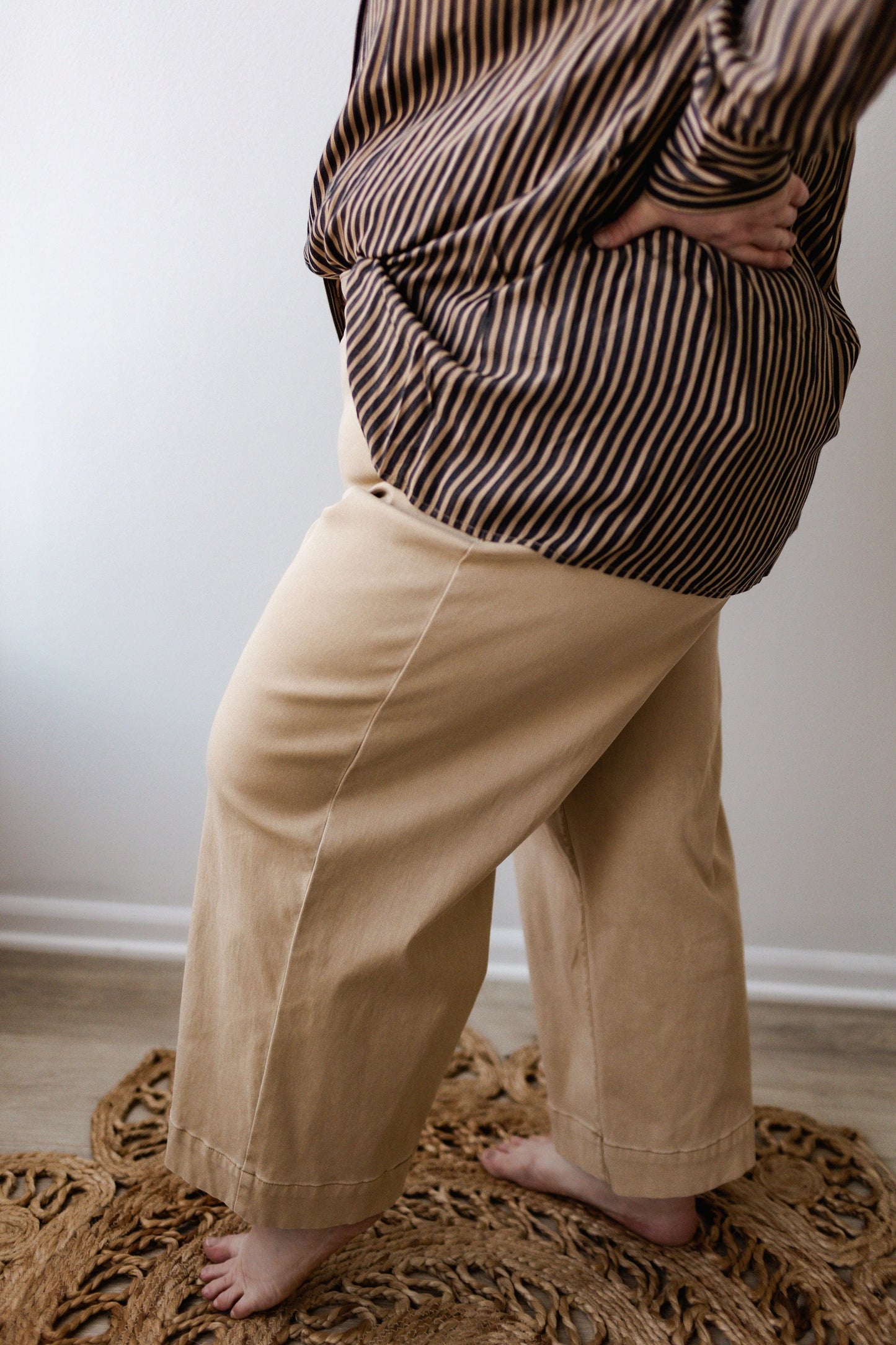 A person wearing a brown striped shirt paired with the Spanx® Stretch Twill Cropped Wide Leg Pant in Almond, standing barefoot on a woven rug.