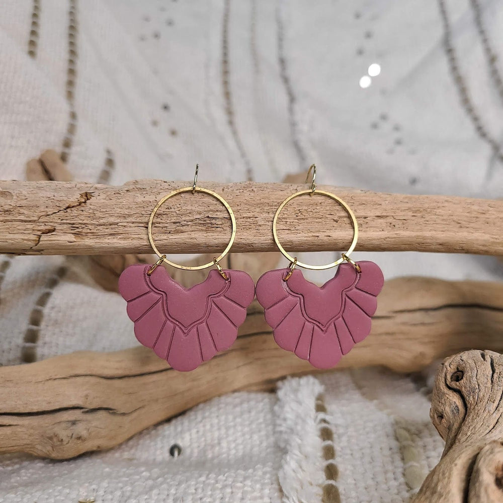 Antique rose fan-shaped earrings with brass hoops hang elegantly on a piece of driftwood against a textured fabric background.