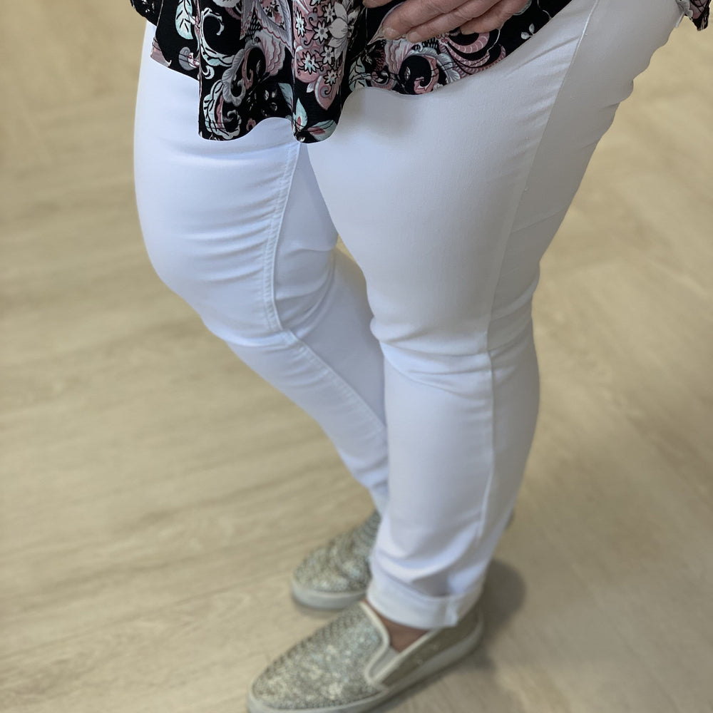 An individual exuding an air of luxury stands on a light-colored wooden floor, hands resting on their hips, dressed in Love Marlow's WHITE DENIM STRETCH SKINNY jeans, a patterned top, and sparkly slip-on shoes.