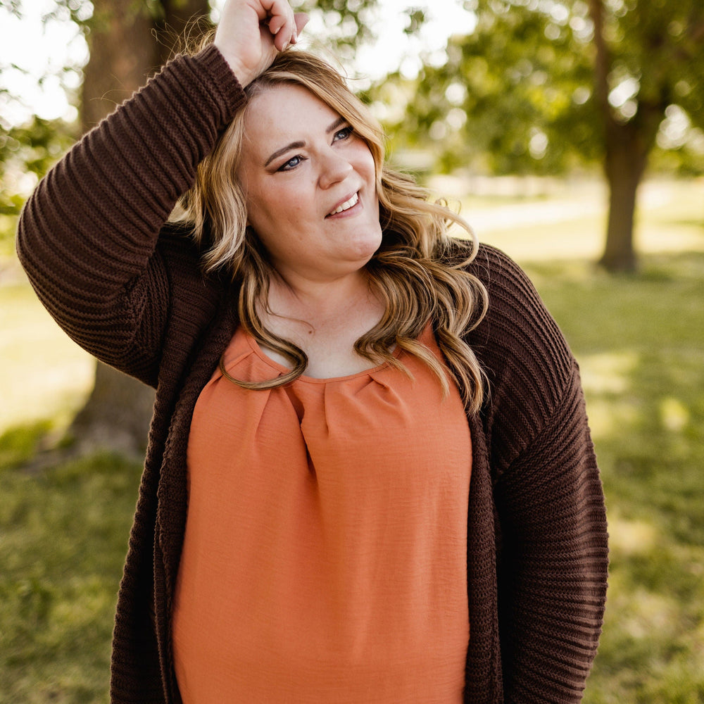 A woman stands outdoors, dressed in a Love Marlow Woven Pleated Neckline Tank in Spiced Pumpkin and a brown cardigan, with her hand raised to adjust her hair.