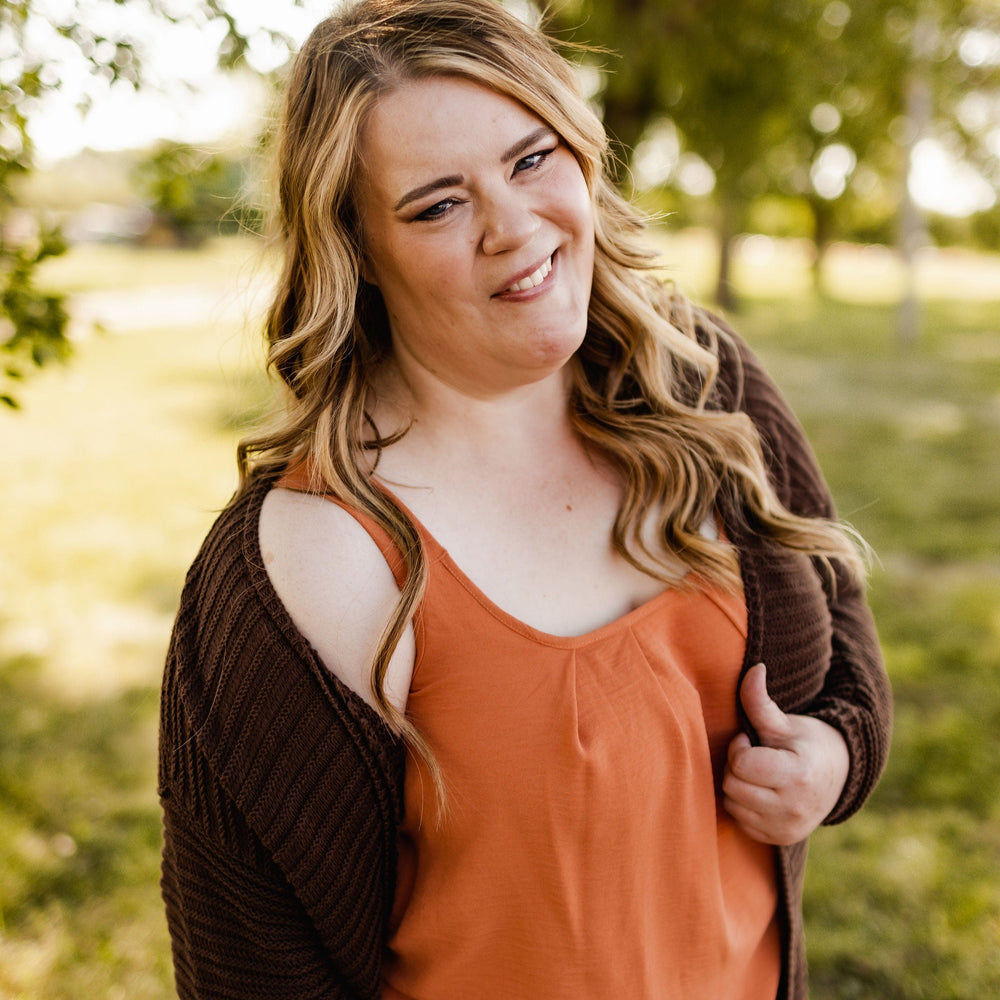 A woman with long wavy hair stands outside in a grassy area, wearing the WOVEN PLEATED NECKLINE TANK IN SPICED PUMPKIN by Love Marlow and a brown cardigan, smiling at the camera.