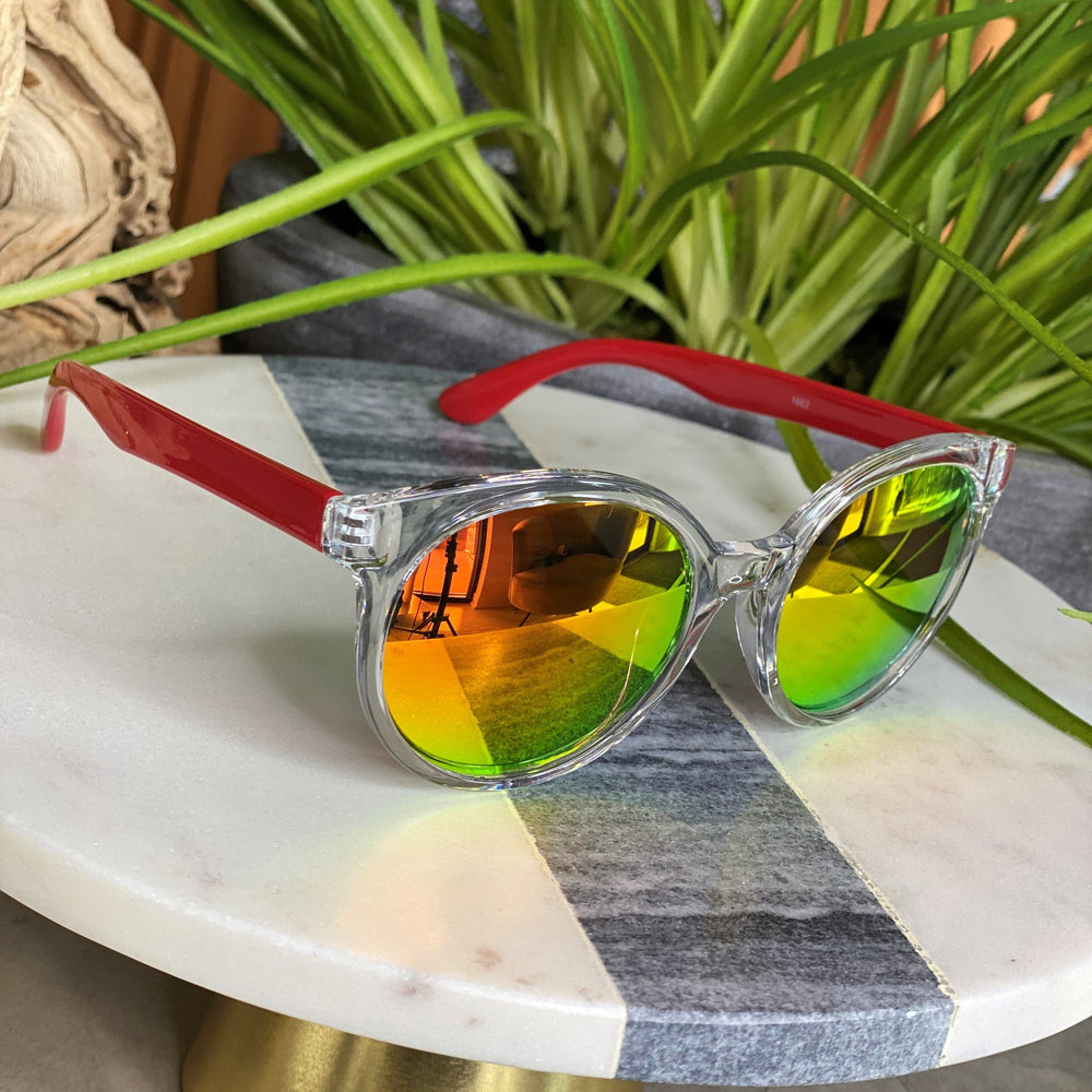 The BARBADOS SUNGLASSES IN RED by Love Marlow, featuring clear frames, red arms, and reflective yellow-green lenses, are showcased on a round marble tabletop with green plants in the background. Providing essential UVA/UVB protection, these stylish sunglasses are ideal for any sunny day in Barbados.