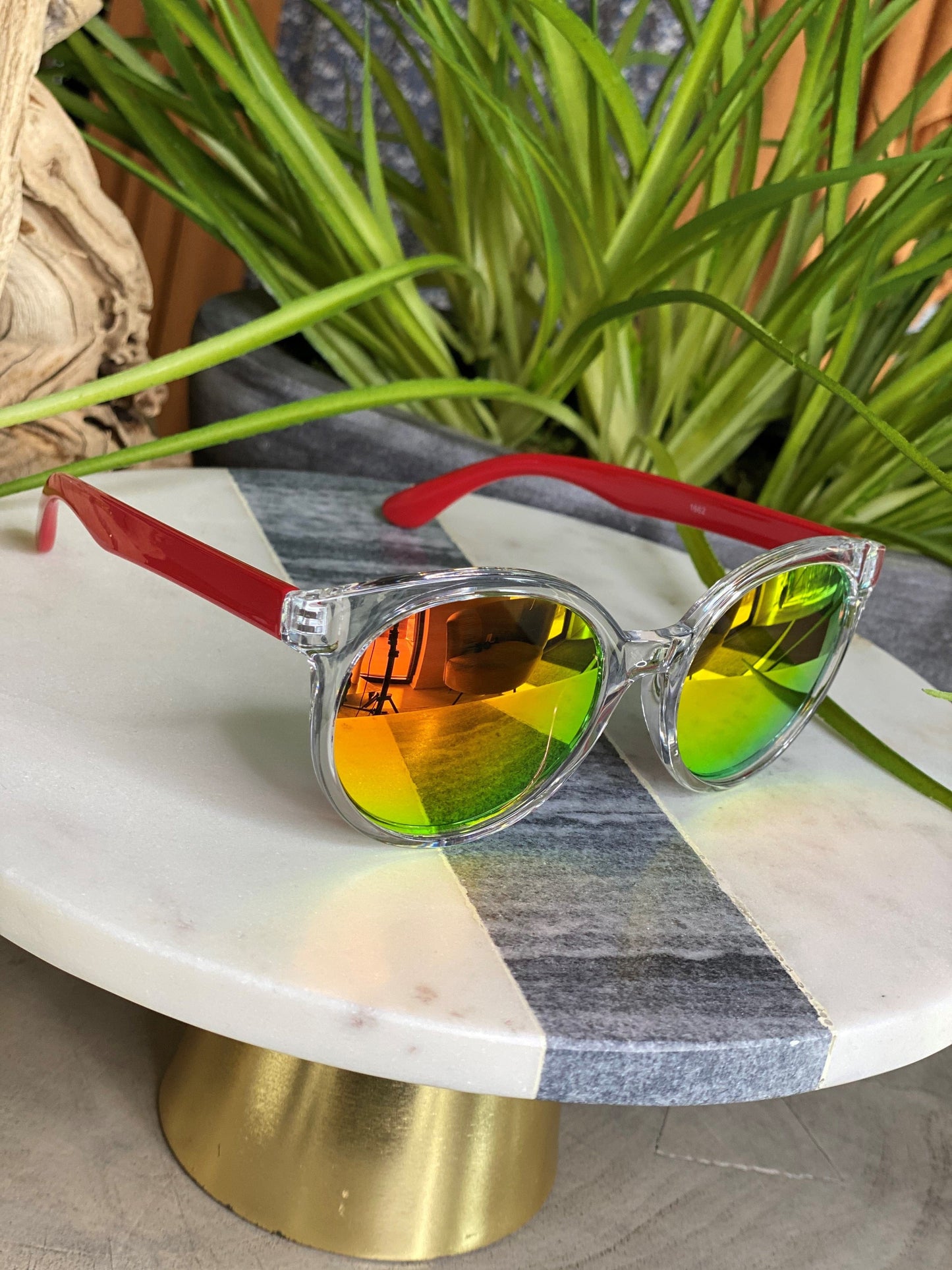 The BARBADOS SUNGLASSES IN RED by Love Marlow, featuring clear frames, red arms, and reflective yellow-green lenses, are showcased on a round marble tabletop with green plants in the background. Providing essential UVA/UVB protection, these stylish sunglasses are ideal for any sunny day in Barbados.