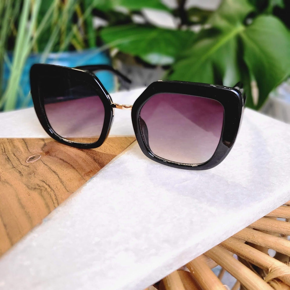 A pair of large, square black sunglasses with gradient lenses exudes 70s vibes as they're displayed on a wooden and marble surface, with green leaves blurred in the background, offering full UVA/UVB protection.