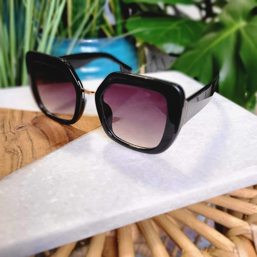 Black square sunglasses with gradient lenses exude 70s vibes as they rest on a white marble surface, surrounded by green plants and a wooden chair. Ideal for those sunny days, they offer full UVA/UVB protection while maintaining timeless style.