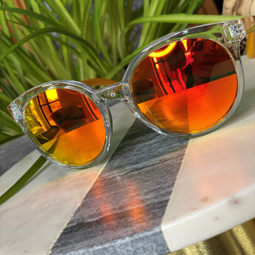 Clear-framed sunglasses with orange mirrored lenses sit stylishly on a marble table, boasting UVA/UVB protection. Green plants create a vibrant backdrop, and with free shipping available, they're both a smart and chic choice.