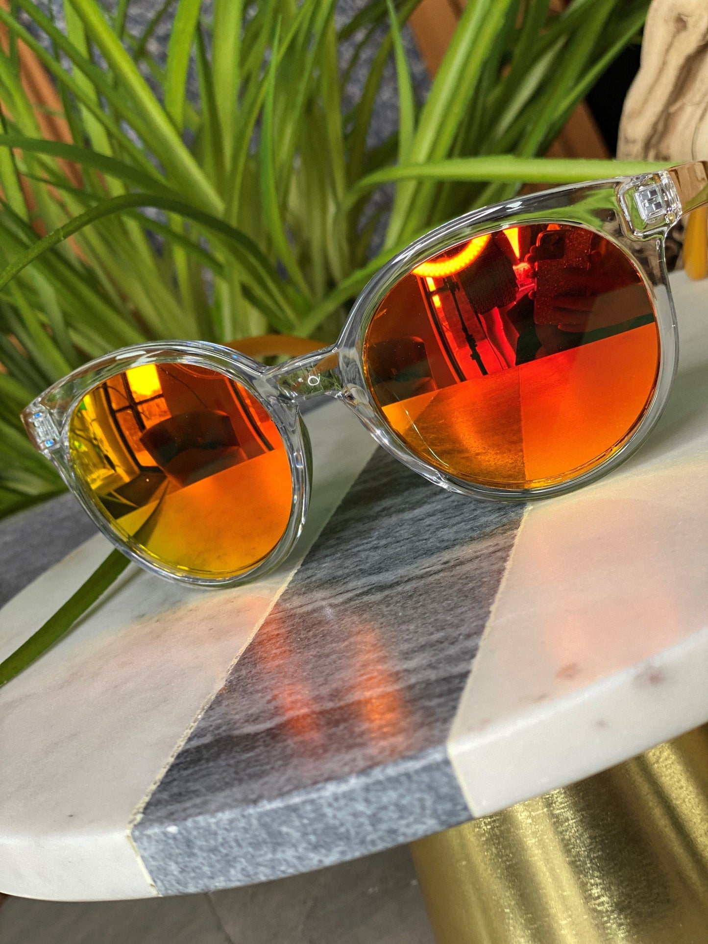 Clear-framed sunglasses with orange mirrored lenses sit stylishly on a marble table, boasting UVA/UVB protection. Green plants create a vibrant backdrop, and with free shipping available, they're both a smart and chic choice.