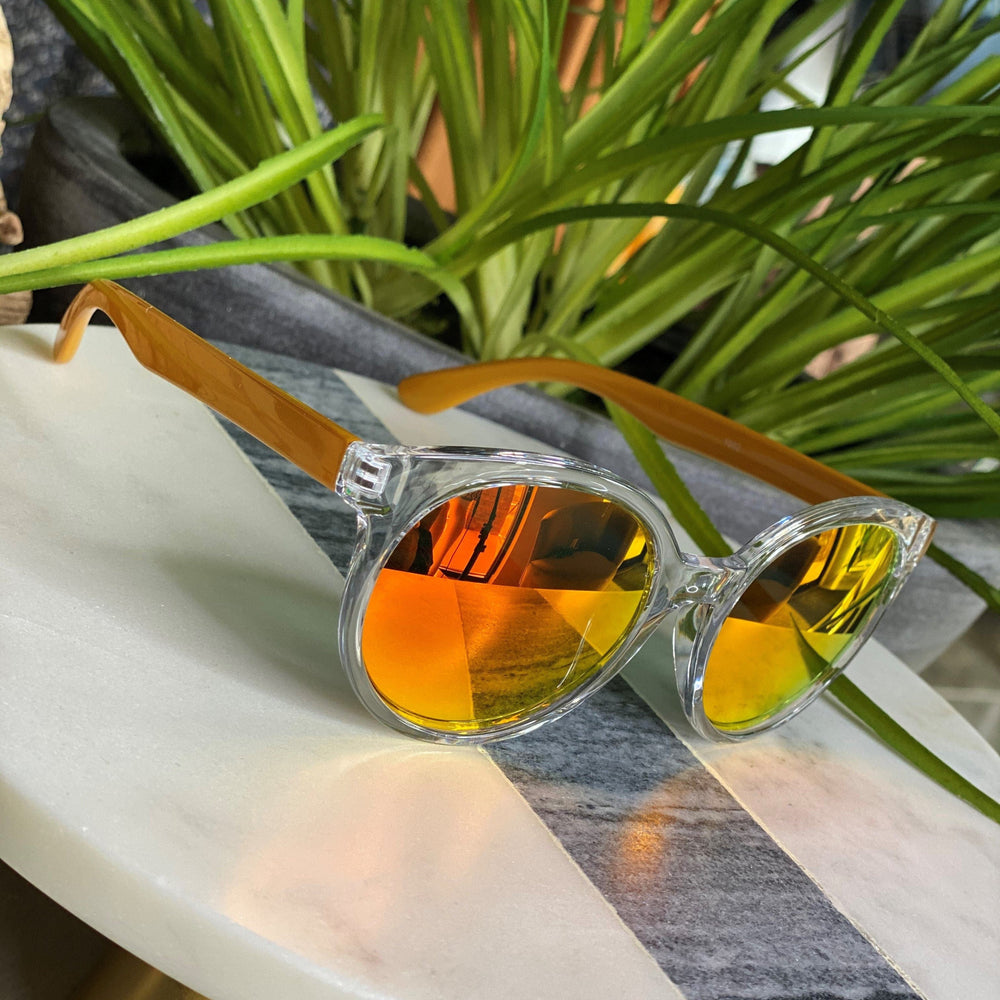 Transparent sunglasses with orange-tinted lenses, offering UVA/UVB protection, rest on a round table with green plants in the background.