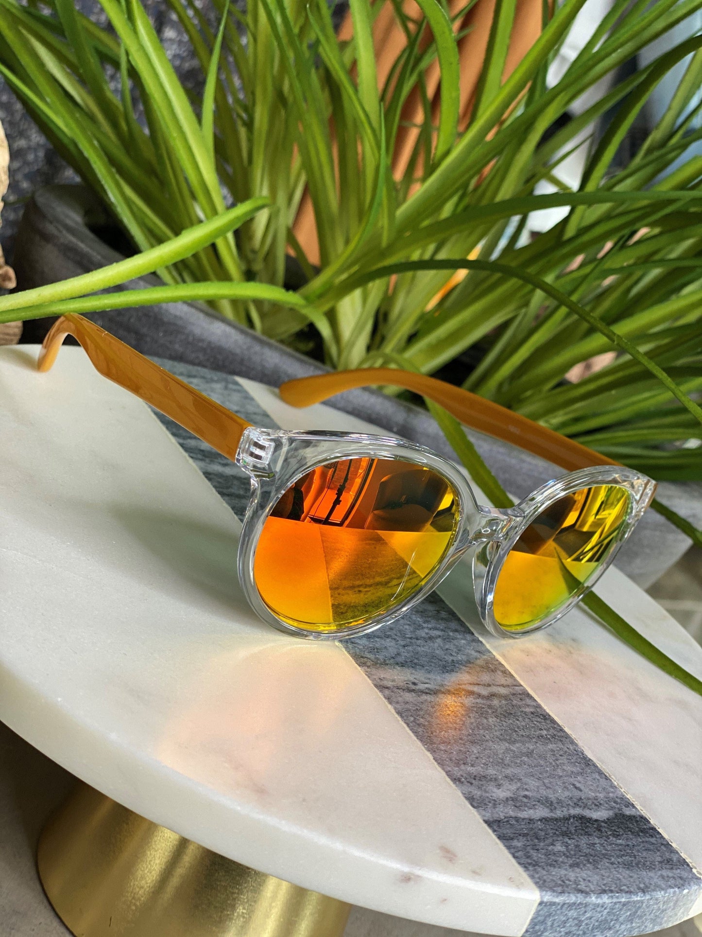 Transparent sunglasses with orange-tinted lenses, offering UVA/UVB protection, rest on a round table with green plants in the background.