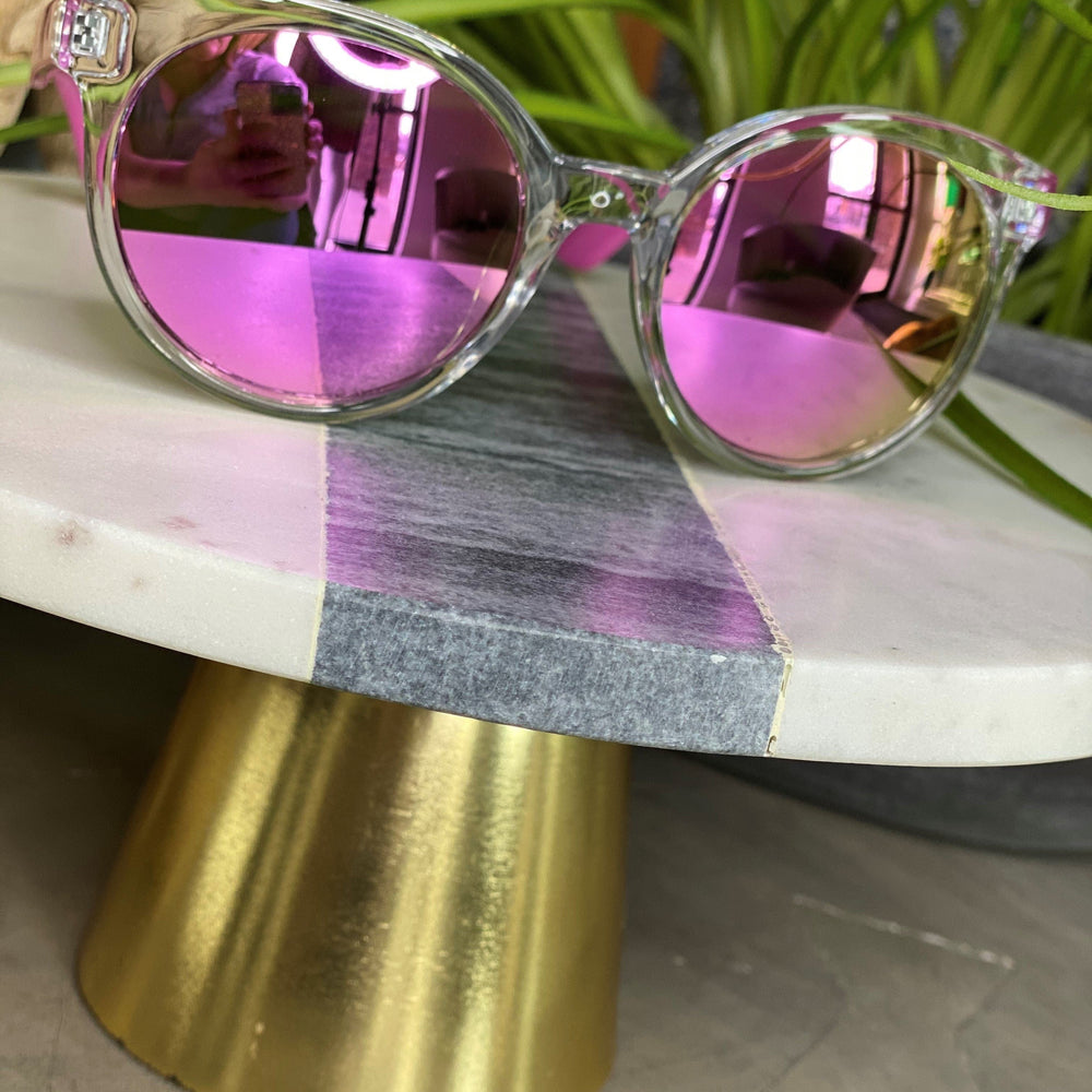 Clear-framed Love Marlow BARBADOS SUNGLASSES IN PINK are placed on a round marble table with a gold base. Green plant leaves are visible in the background, adding a touch of nature to the scene. With built-in UVA/UVB protection, these stylish sunglasses ensure your eyes stay safe while looking chic.
