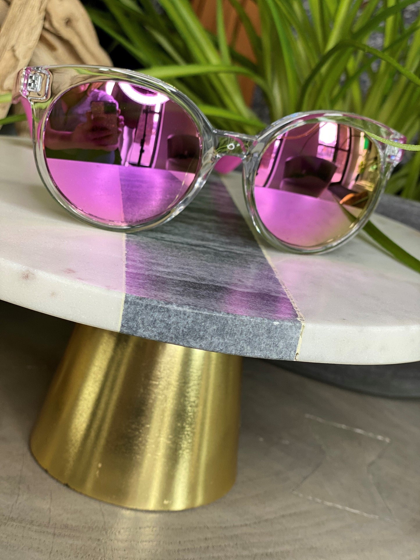 Clear-framed Love Marlow BARBADOS SUNGLASSES IN PINK are placed on a round marble table with a gold base. Green plant leaves are visible in the background, adding a touch of nature to the scene. With built-in UVA/UVB protection, these stylish sunglasses ensure your eyes stay safe while looking chic.