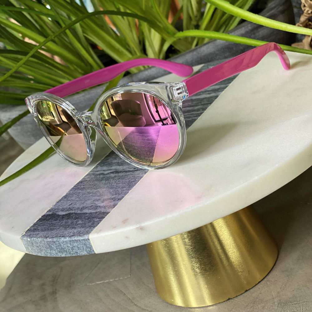 Clear-framed BARBADOS SUNGLASSES IN PINK by Love Marlow with pink arms and reflective lenses displayed on a white and gray marble stand with a golden base, next to green plants. These stylish sunglasses not only offer UVA/UVB protection but also come with free shipping.
