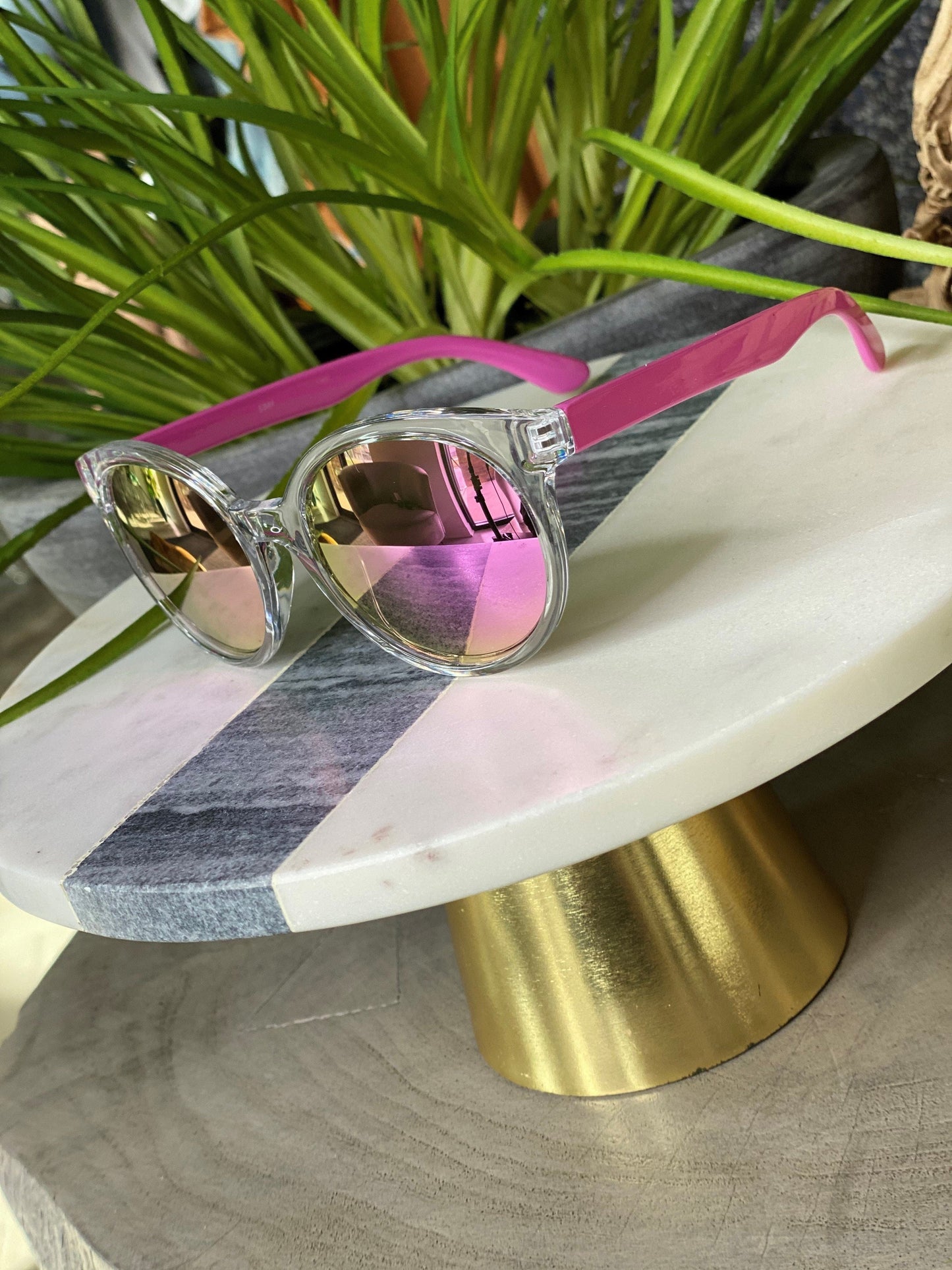 Clear-framed BARBADOS SUNGLASSES IN PINK by Love Marlow with pink arms and reflective lenses displayed on a white and gray marble stand with a golden base, next to green plants. These stylish sunglasses not only offer UVA/UVB protection but also come with free shipping.