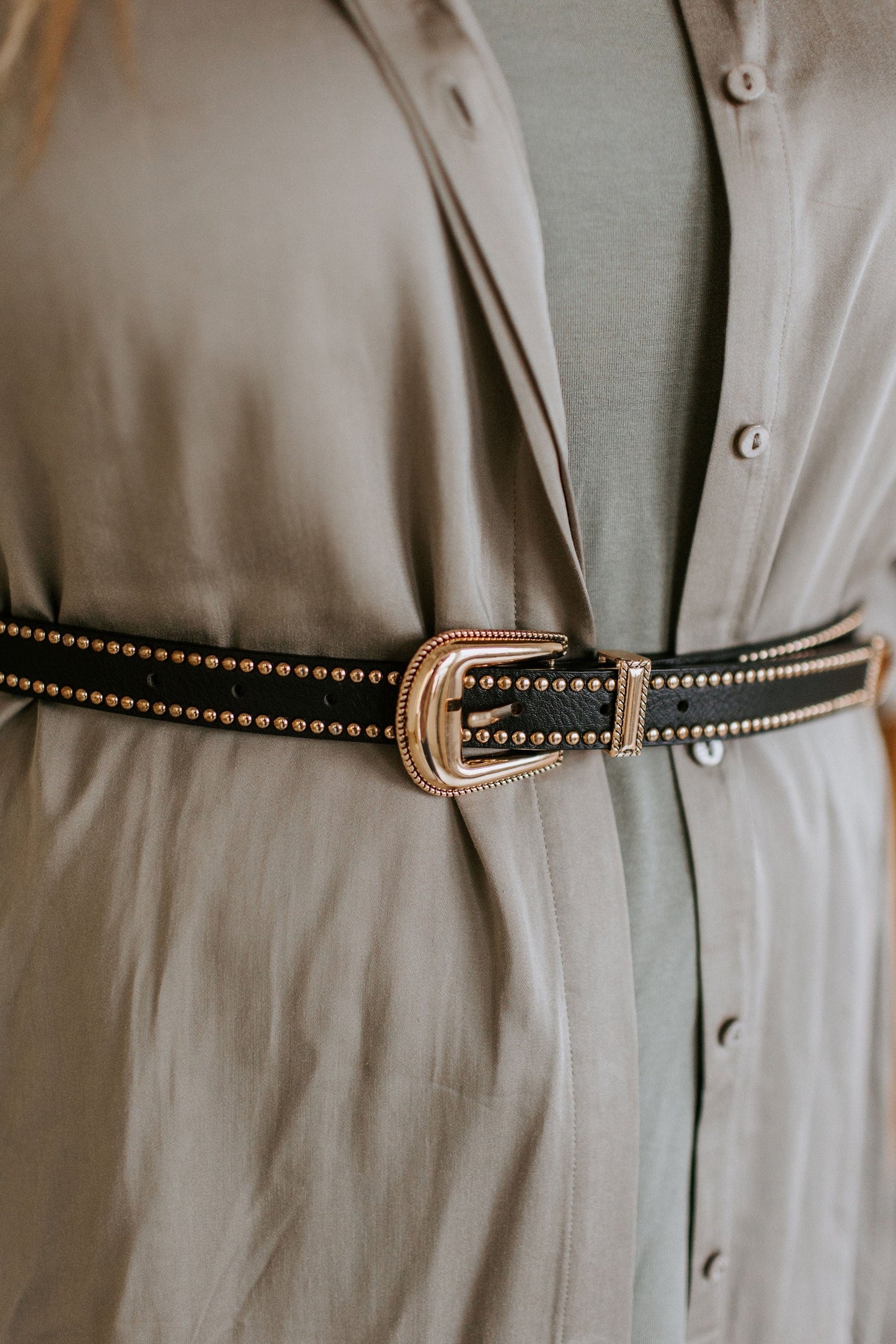 Close-up of a person wearing a beige blouse with a faux leather belt, waist-defining with gold-tone grommets, fastened at the waist.