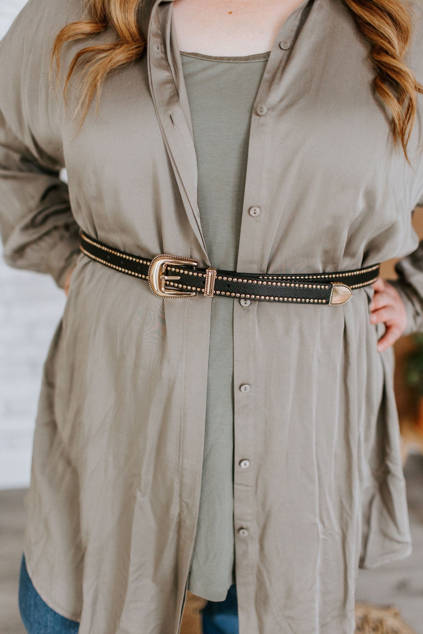 A person wearing a loose, light grey shirt with a waist-defining faux leather belt featuring gold-tone grommets stands with hands on hips. Their hair is down and they are wearing jeans.