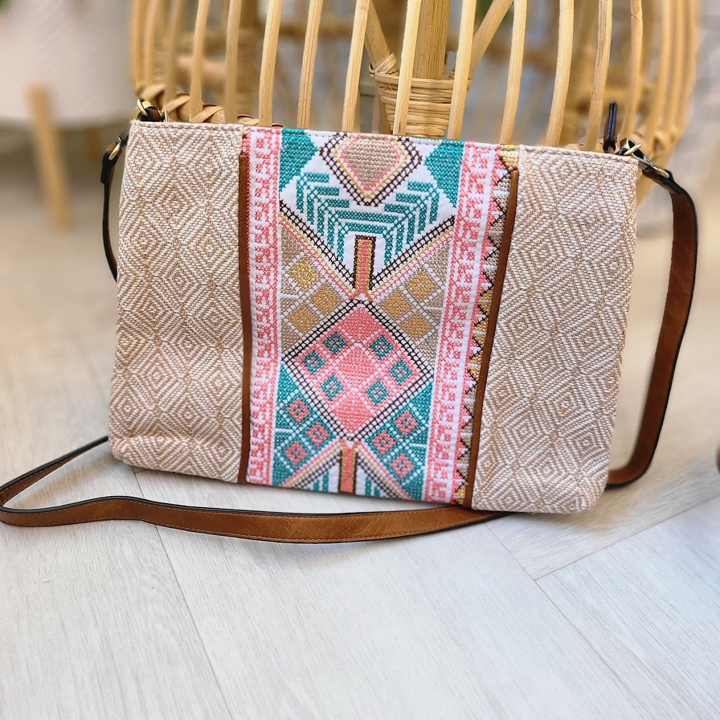 A boho-chic crossbody purse with a geometric pattern in pink, teal, and peach, crafted from vegan faux leather and featuring a brown strap, sits artfully on a light wood floor.