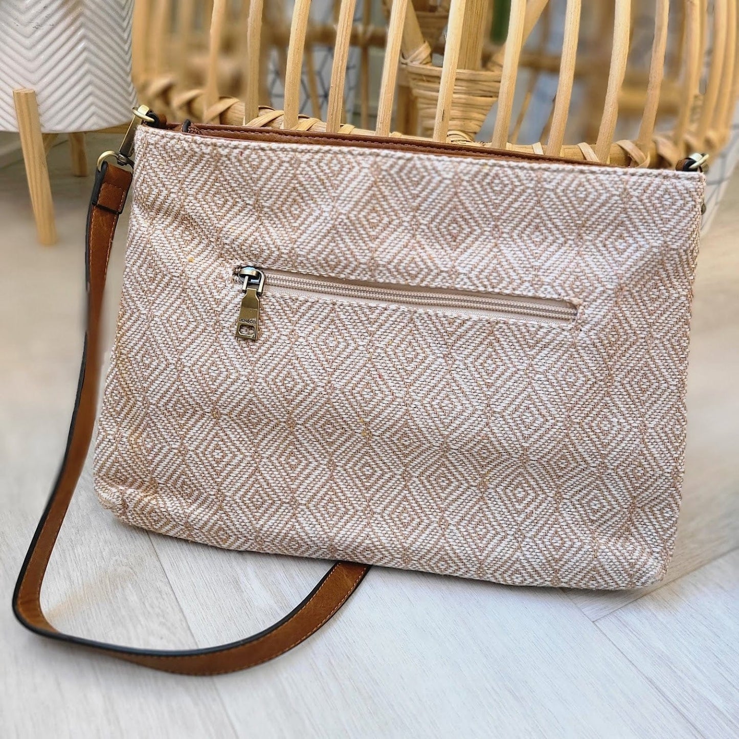 A beige patterned crossbody purse crafted from vegan faux leather features a front zipper pocket and a brown strap, elegantly placed on a light wooden floor.