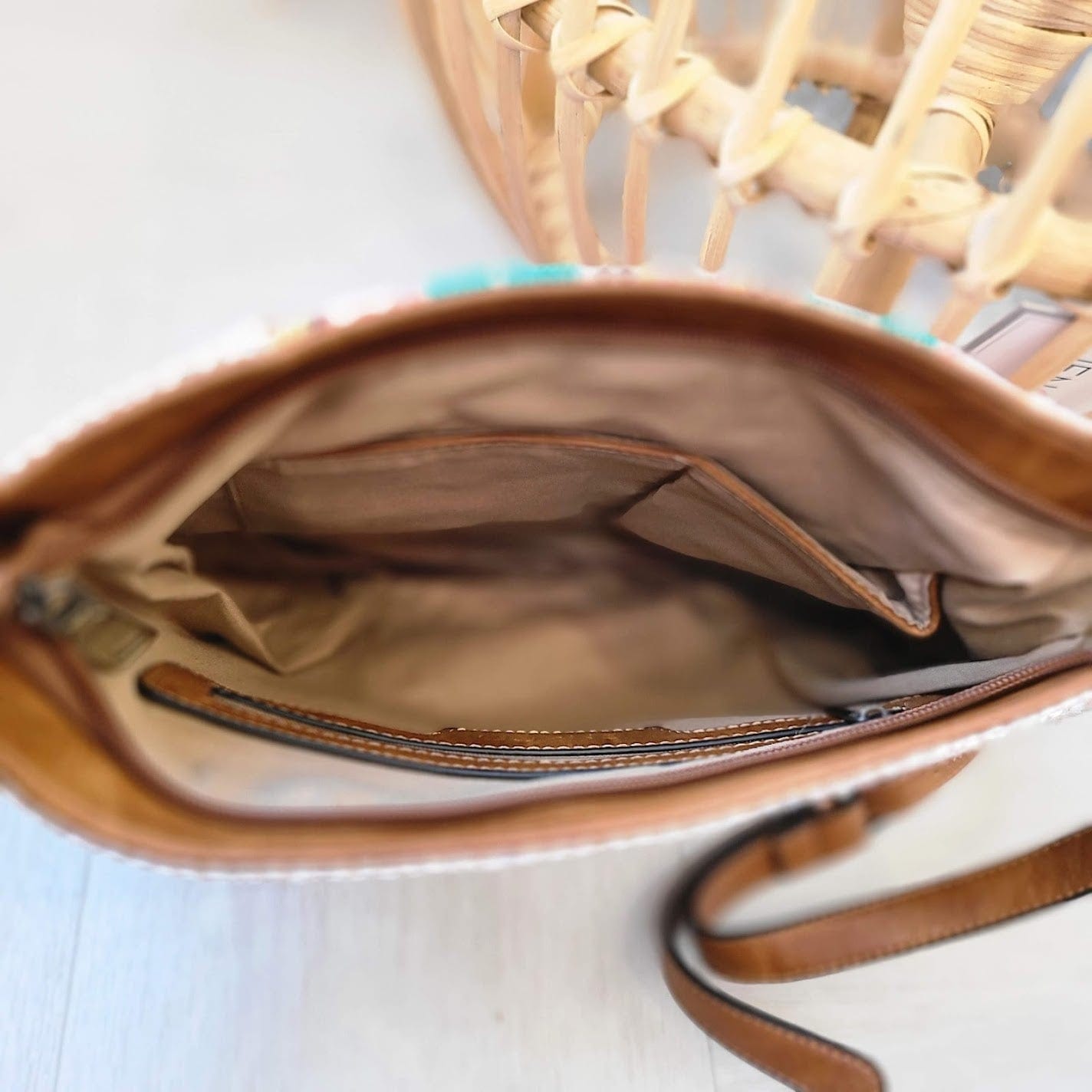 An open brown handbag with a beige interior, crafted from vegan faux leather, sits elegantly on a light surface next to a wicker chair.