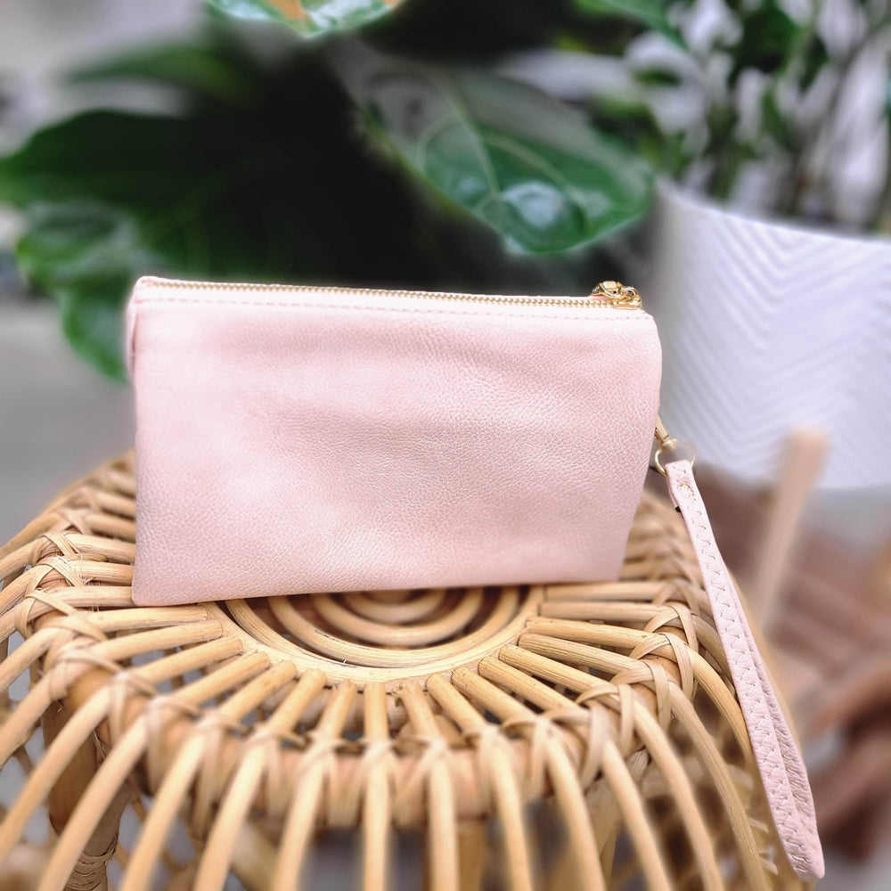 A light pink faux leather clutch with a detachable strap rests gracefully on a woven rattan stool, surrounded by lush green plants.