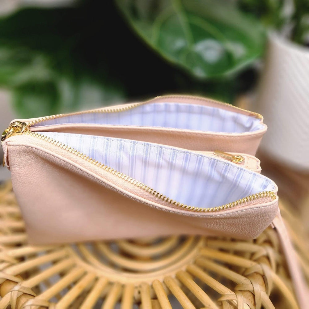 A beige faux leather bag with gold zippers is open, revealing a striped fabric lining. It sits on a woven surface with green plants in the background, and its detachable strap offers versatility.