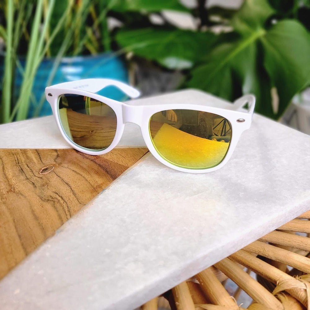 White sunglasses with a 90s frame and yellow-tinted lenses rest on a white and wooden surface, surrounded by green plants.