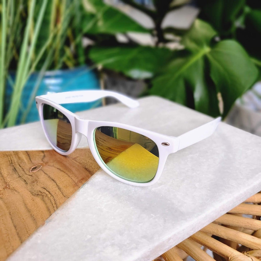 White sunglasses with a 90s frame and reflective lenses rest stylishly on a wooden and marble surface, surrounded by lush green plants.