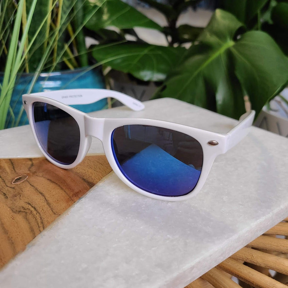 White sunglasses with a 90s frame and blue reflective lenses rest on a wooden and marble surface, surrounded by green leaves. These stylish shades offer essential UVA/UVB protection, making them both trendy and functional for any sunny day.