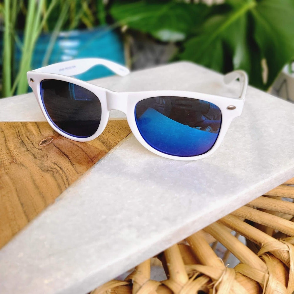 White sunglasses with a 90s frame and blue lenses rest on a wooden and marble surface amidst greenery, offering stylish UVA/UVB protection.
