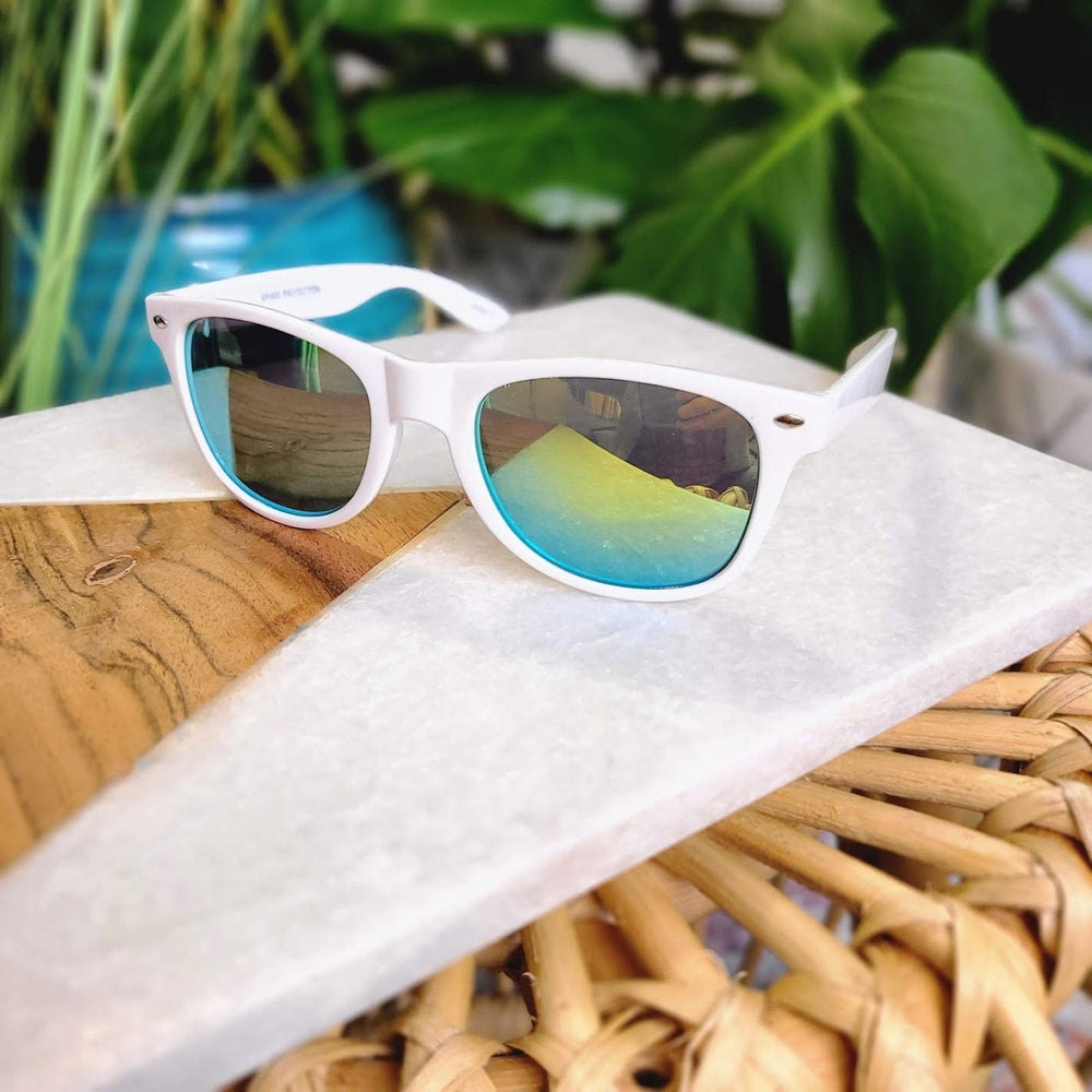 White sunglasses with a 90s frame and reflective blue lenses rest on a marble and wooden table, providing UVA/UVB protection against blurred green plants in the background.