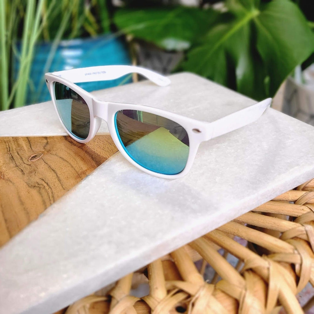 White sunglasses with a sleek 90s frame and blue-tinted lenses rest on a marble and wood surface, surrounded by greenery. These stylish shades offer full UVA/UVB protection for your eyes.