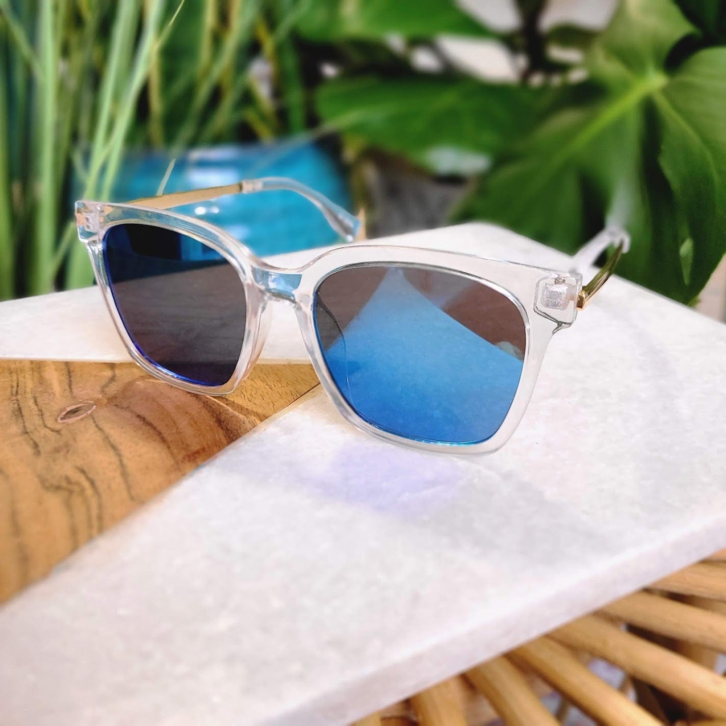 Clear-framed sunglasses with blue mirror lenses offering UVA/UVB protection rest on a wooden and white table, surrounded by greenery in the background.