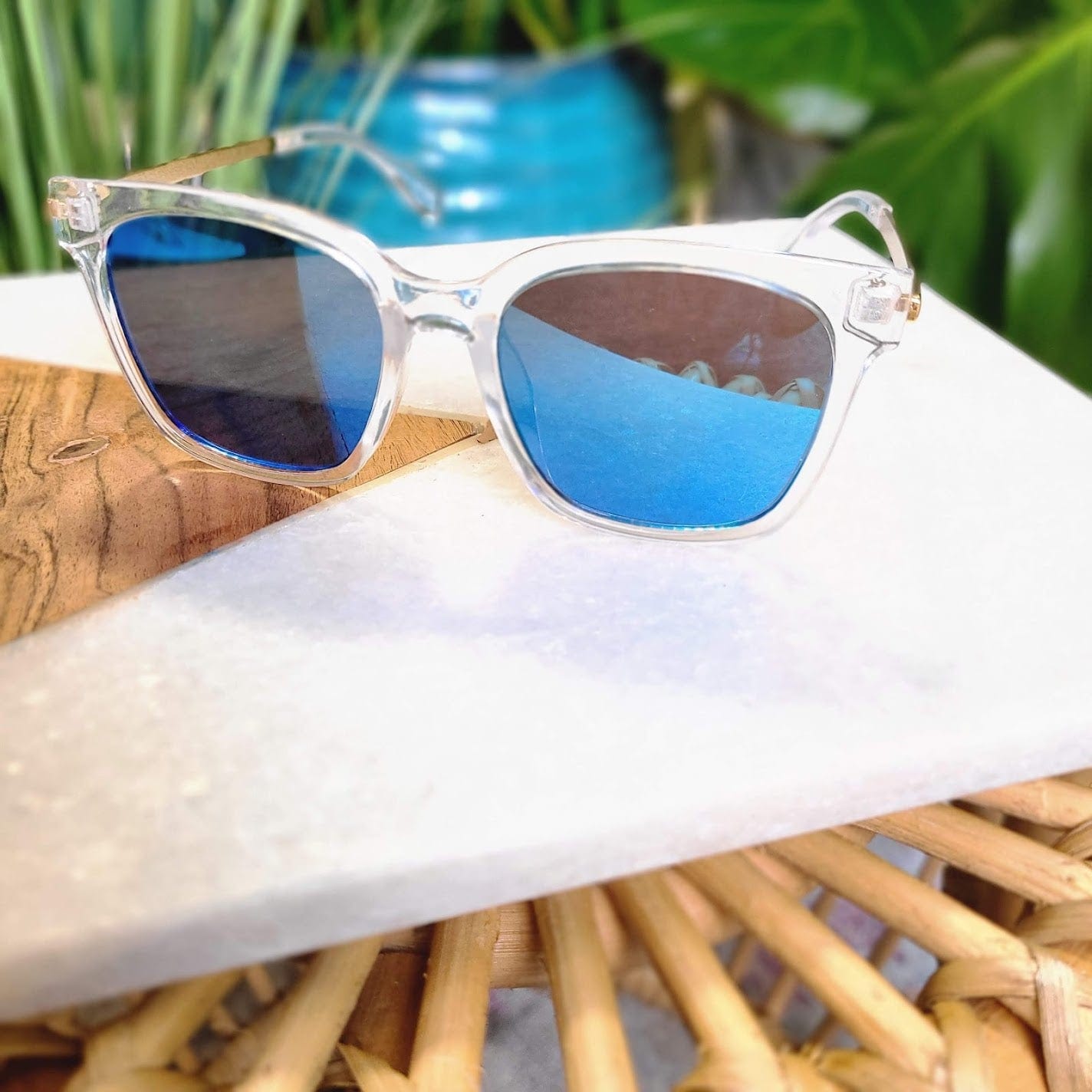 Clear-framed sunglasses with blue mirror lenses sit on a marble surface, surrounded by wooden and wicker textures. Featuring full UVA/UVB protection, they are perfect for sunny days, with lush greenery providing a vibrant backdrop.