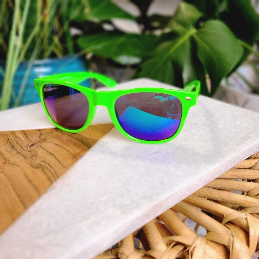 Bright green 90s frame sunglasses with blue-tinted lenses rest on a geometric-patterned surface, offering full UVA/UVB protection.