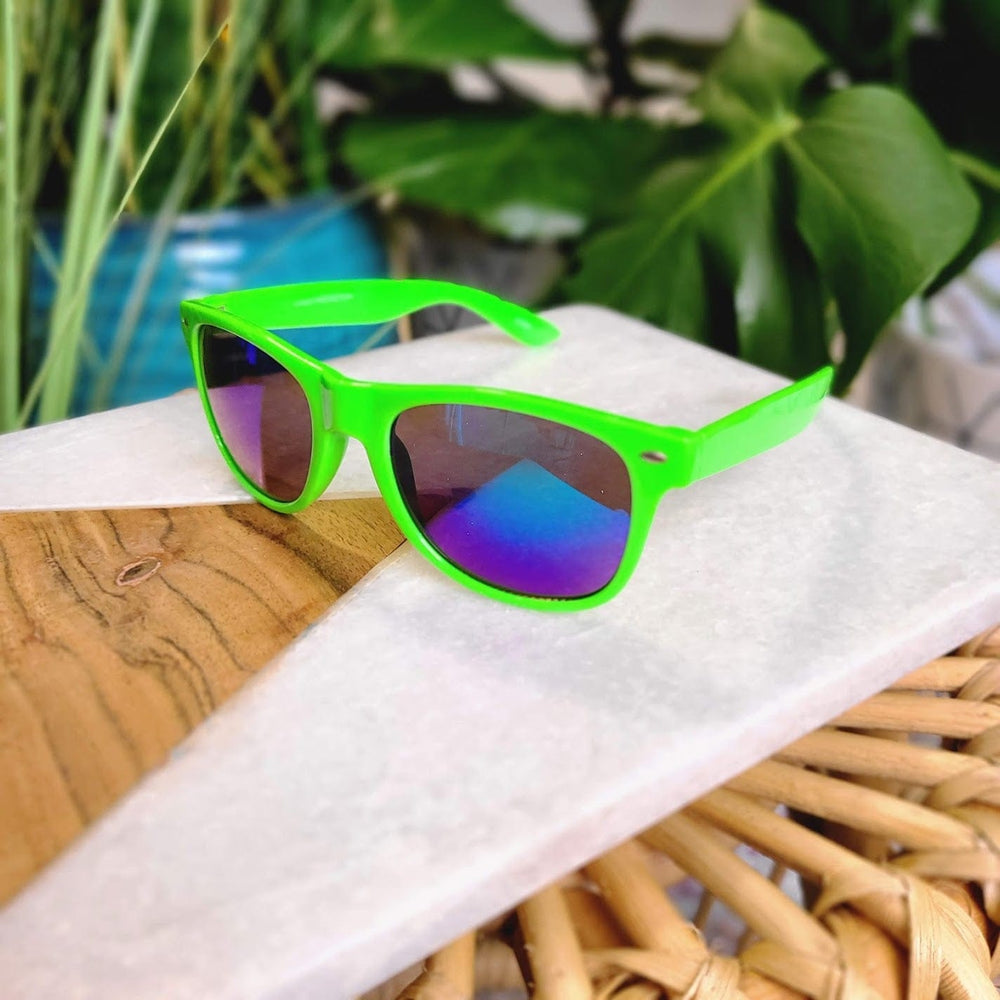 Bright green sunglasses with tinted lenses, offering UVA/UVB protection, rest on a white and wooden surface. Surrounded by lush green plants in the background, their retro 90s frame adds a nostalgic touch to the scene.