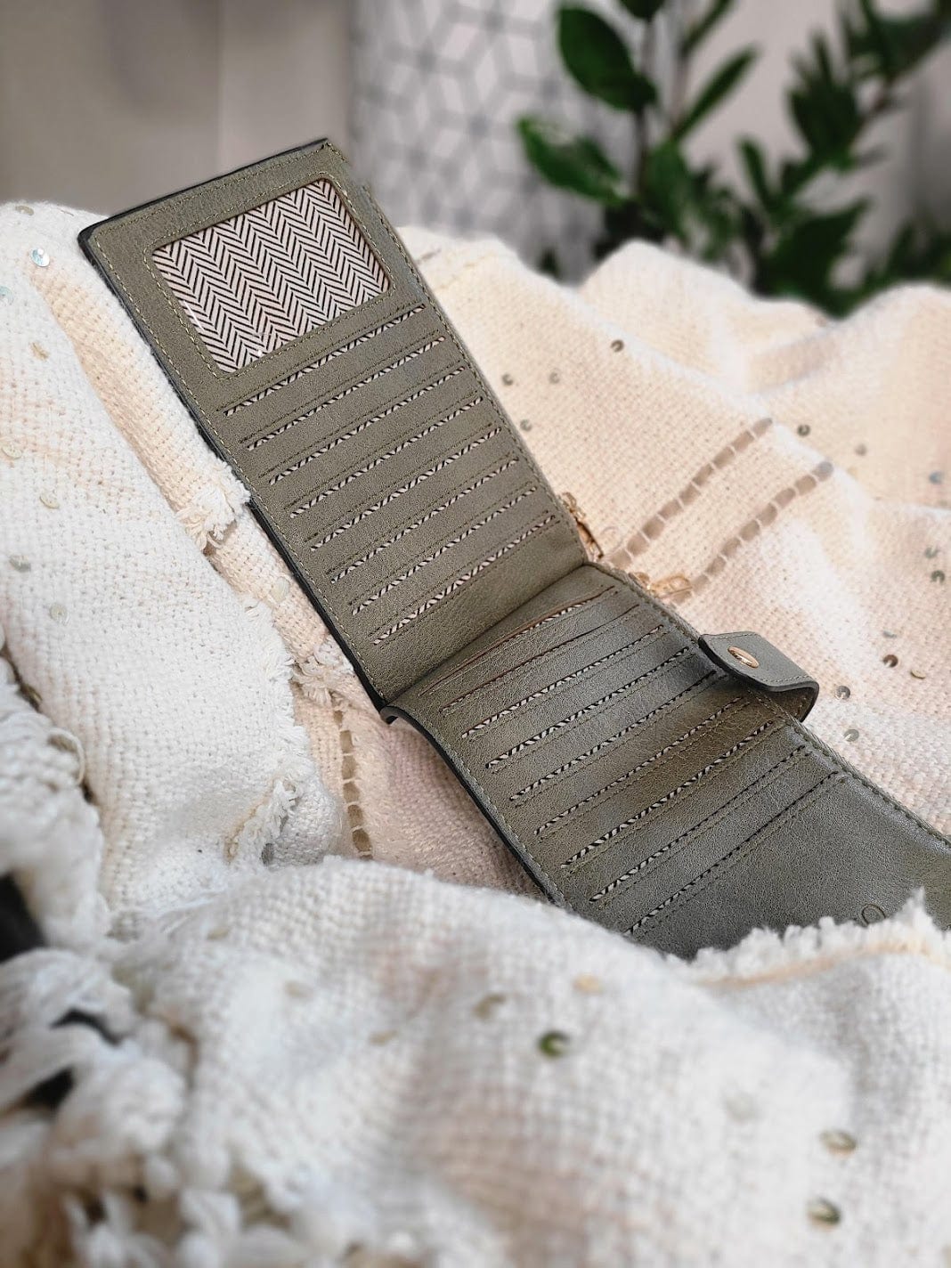 An army green wallet is open, showcasing several card slots with RFID protection. It rests on a textured white fabric, complemented by faint background foliage.