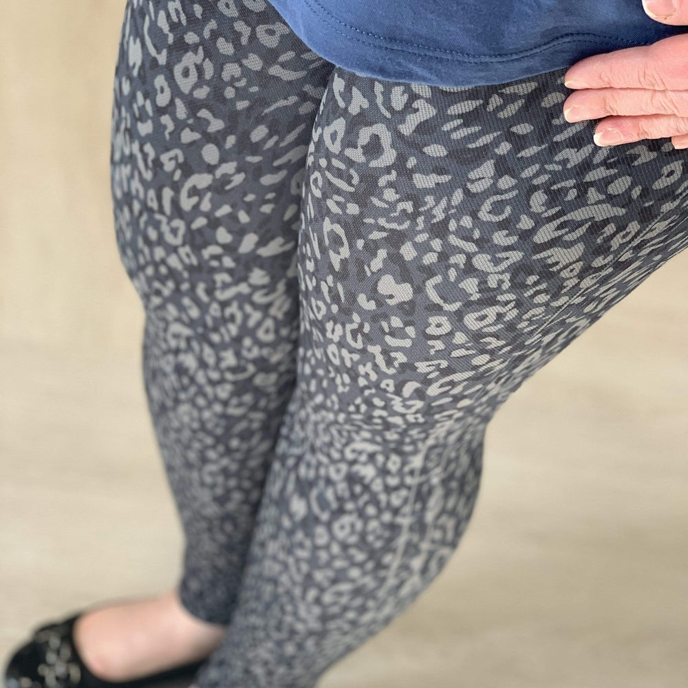A person wearing the Spanx© LOOK AT ME NOW SEAMLESS LEGGING in a gray leopard print, paired with a blue top and black shoes, stands on a light floor, appreciating the shaping support of the double-layer waistband.