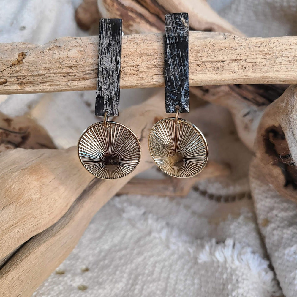 A pair of artistic Cow Horn Earrings with circular metal designs hanging from rectangular black and white marbled posts, displayed on natural wooden branches.